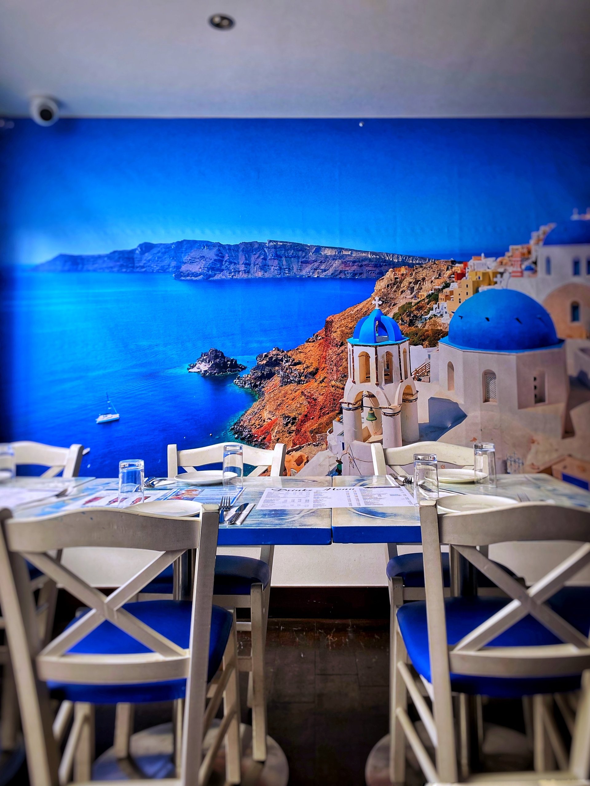 Restaurant interior with white chairs facing a large wall mural of Greek island architecture overlooking blue Mediterranean sea