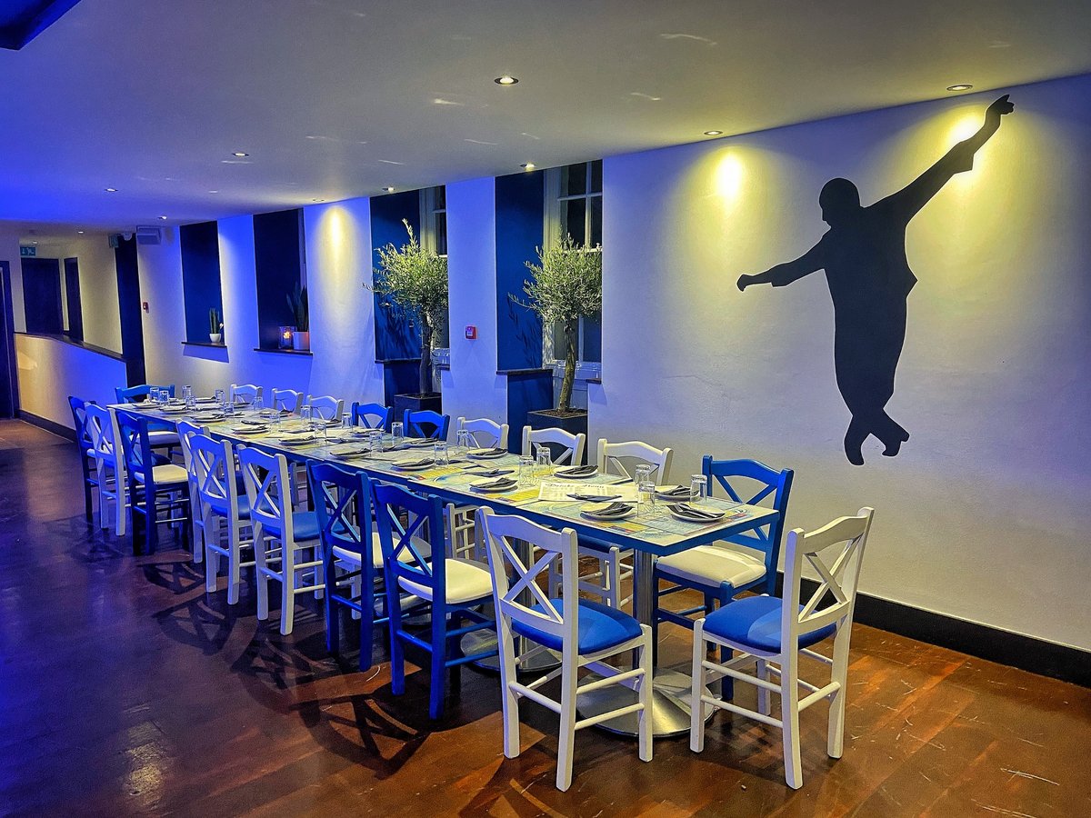 A long table set with place settings, blue and white chairs, and a silhouette figure on the wall at Zorba The Greek