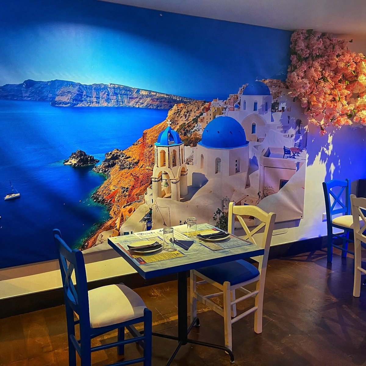 A table set against a large mural of a Greek seaside village with white buildings and blue domes overlooking the sea, at Zorba The Greek